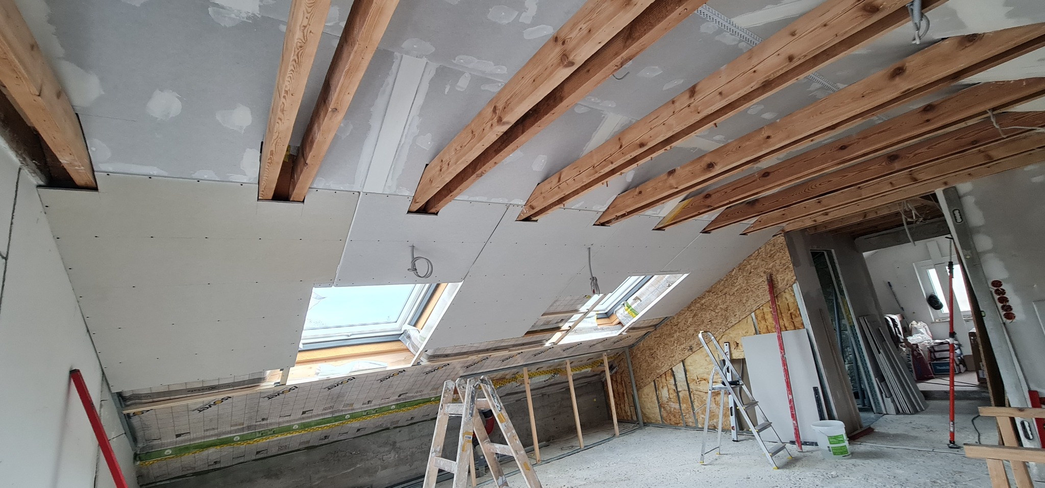 Montaż płyt gipsowo-kartonowych na poddaszu, widoczne drewniane belki stropowe, okna dachowe i drabiny, pomieszczenie w trakcie prac wykończeniowych.