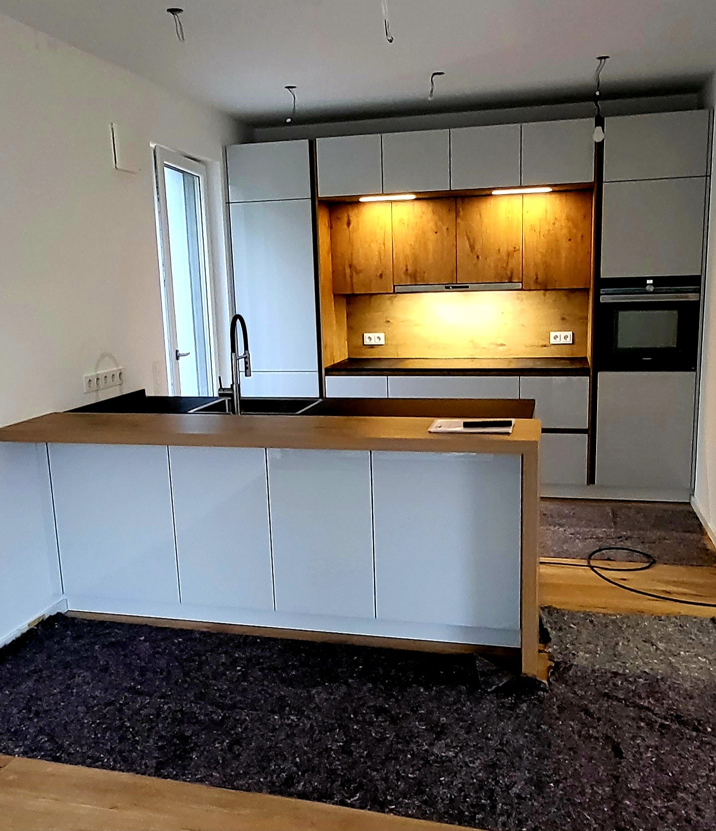 Nowoczesna kuchnia w trakcie montażu: białe szafki z drewnianym blatem, oświetlenie podszafkowe, widoczne niedokończone wykończenia podłogi i kable zwisające z sufitu.