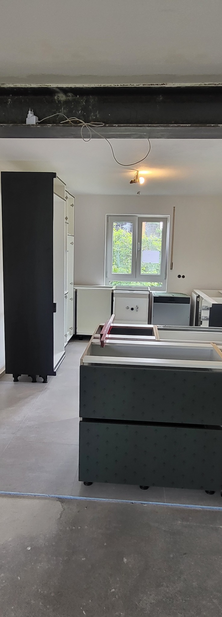 Wnętrze w trakcie montażu zabudowy kuchennej: widoczne szafki dolne z ciemnozielonymi frontami w drobny wzór, białe szafki wiszące, surowy betonowy sufit z widoczną instalacją elektryczną i lampą...