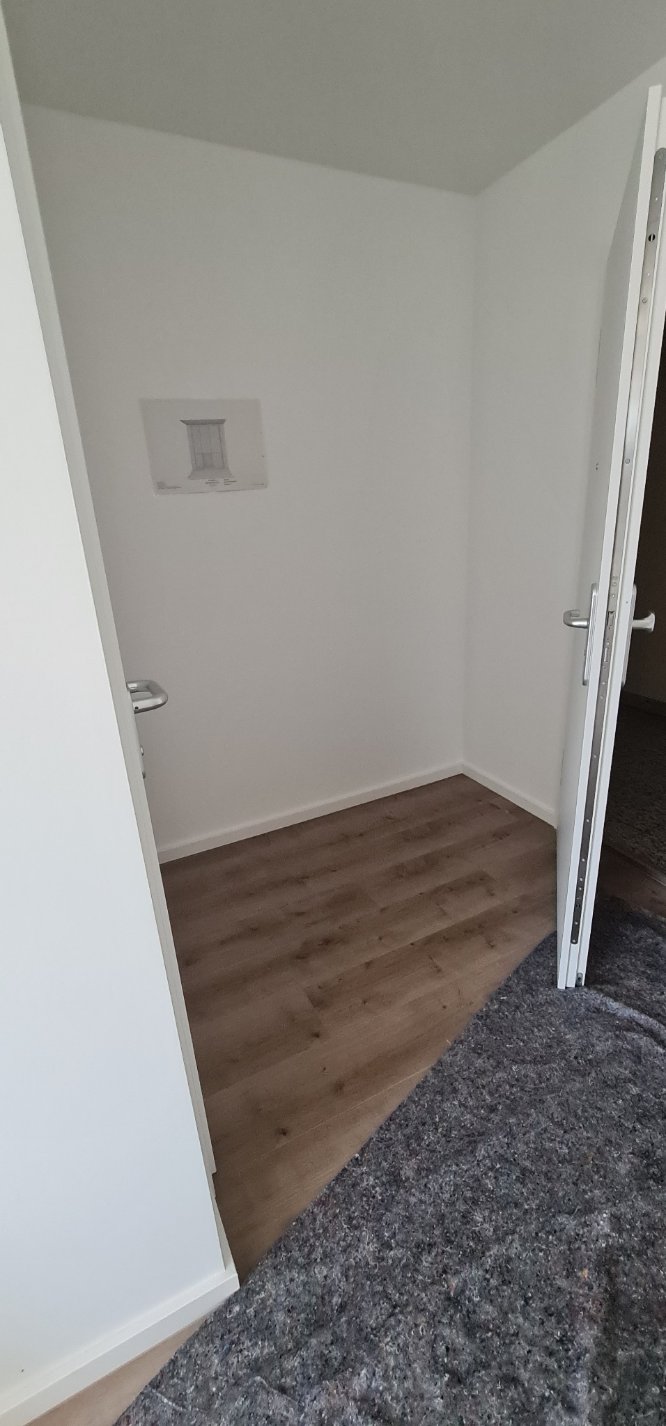 Pusta garderoba z panelami podłogowymi, białe ściany i szkic okna na ścianie, otwarte białe drzwi z srebrną klamką, zabezpieczenie podłogi przed uszkodzeniami.