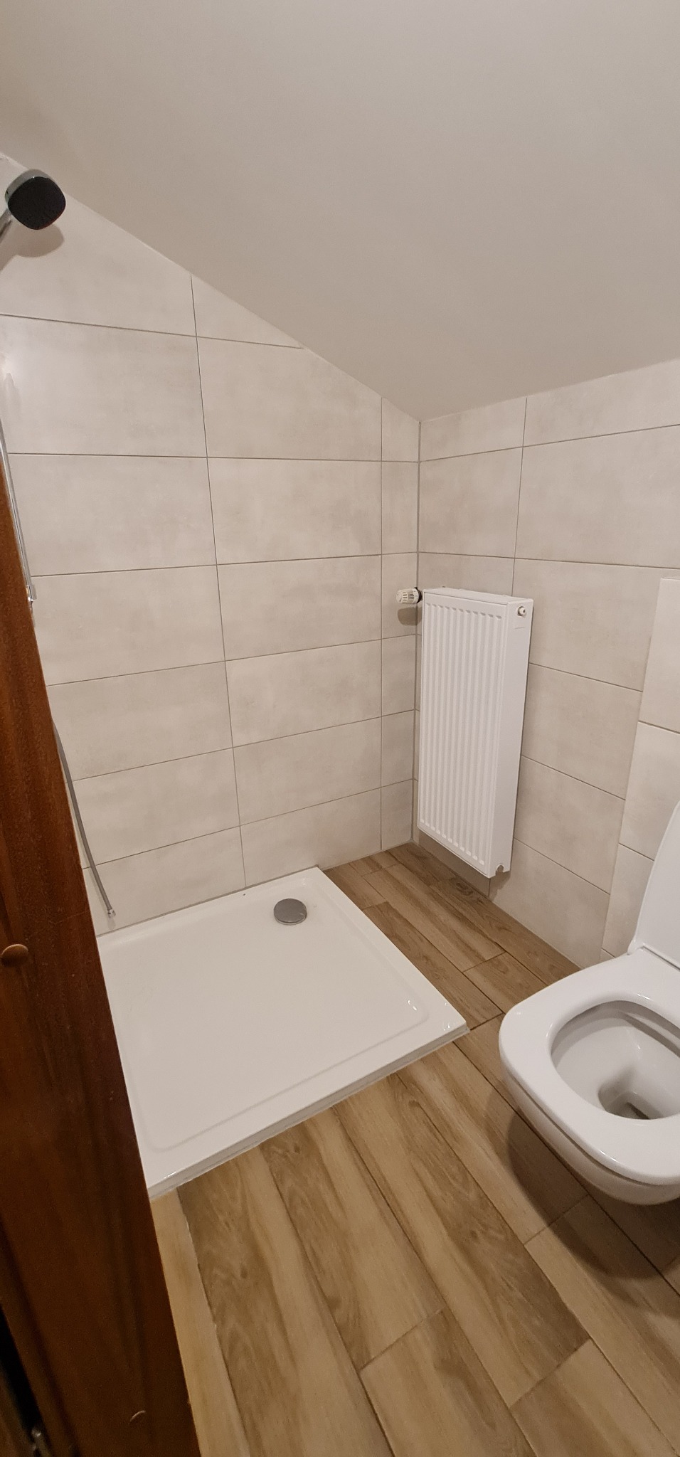 Nowoczesna łazienka z beżowymi płytkami ściennymi, drewnopodobną podłogą, białą kabiną prysznicową, grzejnikiem i toaletą.