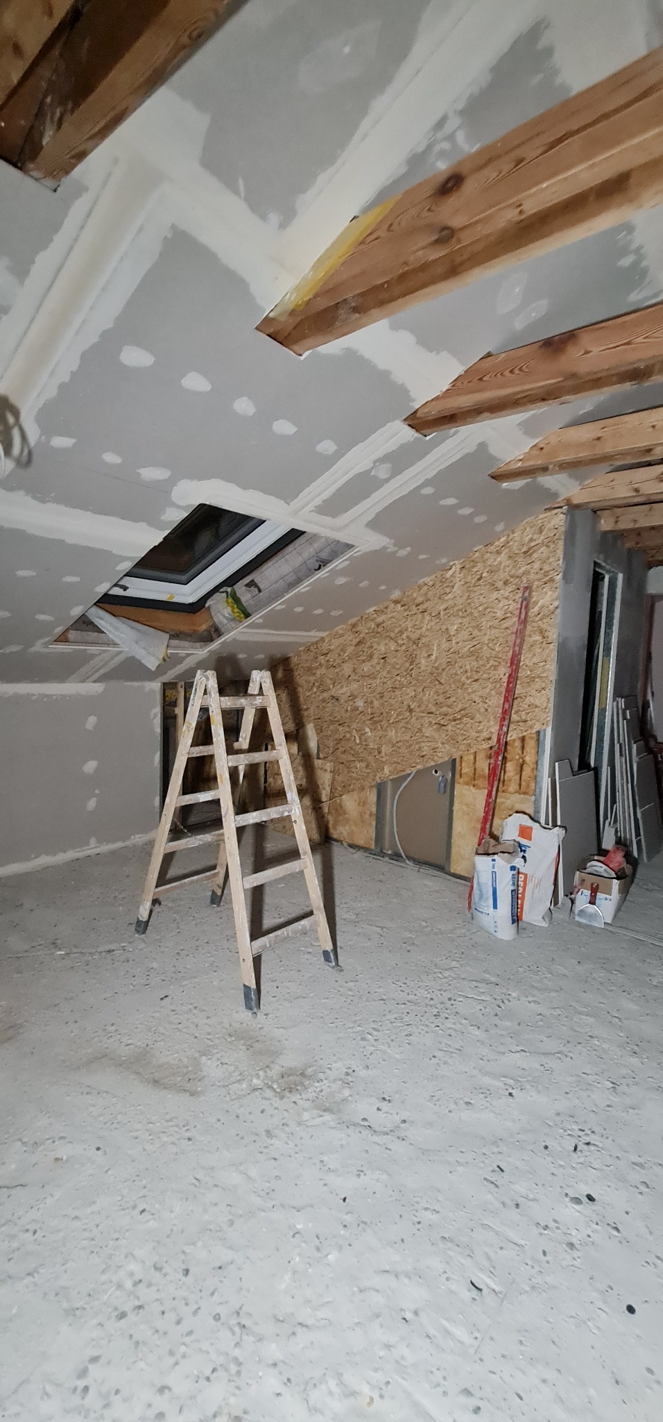 Wnętrze w trakcie remontu: sufit z płyt gipsowo-kartonowych z widocznym otworem na okno dachowe, drewniane elementy konstrukcyjne, drabina, płyty i worki z zaprawą na podłodze.