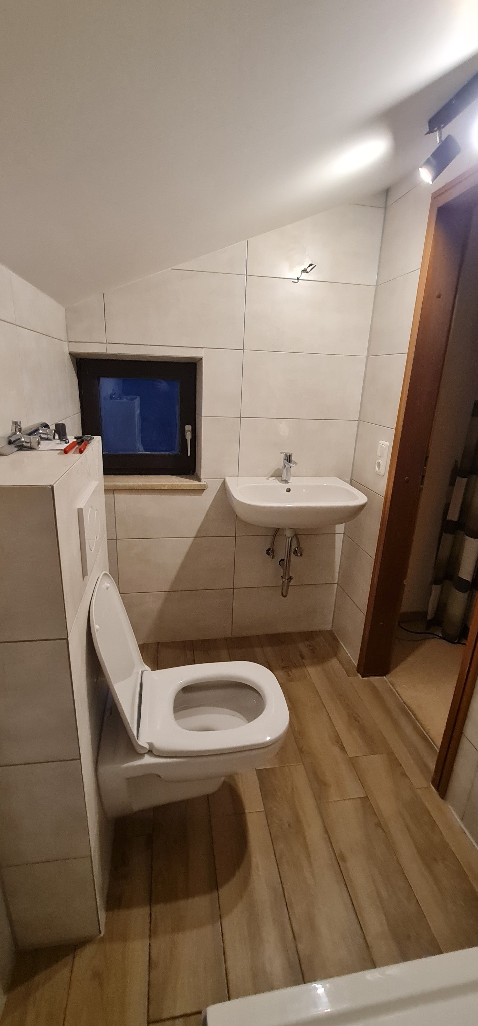 Wnętrze łazienki z wiszącą toaletą z otwartą klapą, umywalką i drewnopodobnymi płytkami podłogowymi. Na blacie obok okna leżą narzędzia hydrauliczne.