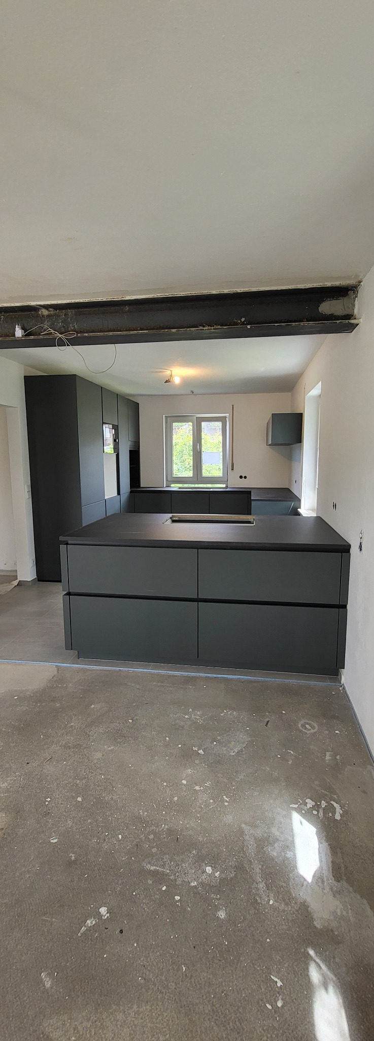 Nowoczesna, szara kuchnia w trakcie wykańczania; widoczna wyspa kuchenna z szufladami, szafki oraz surowy beton na podłodze i metalowa belka stropowa.