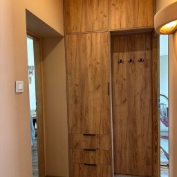 Meble, kuchnie, szafy, stoliki na wymiar. - Wysoka szafa z frontami imitującymi drewno, z trzema szufladami na dole i górną szafką, wkomponowana w przedpokój z wieszakami na ścianie wnęki.