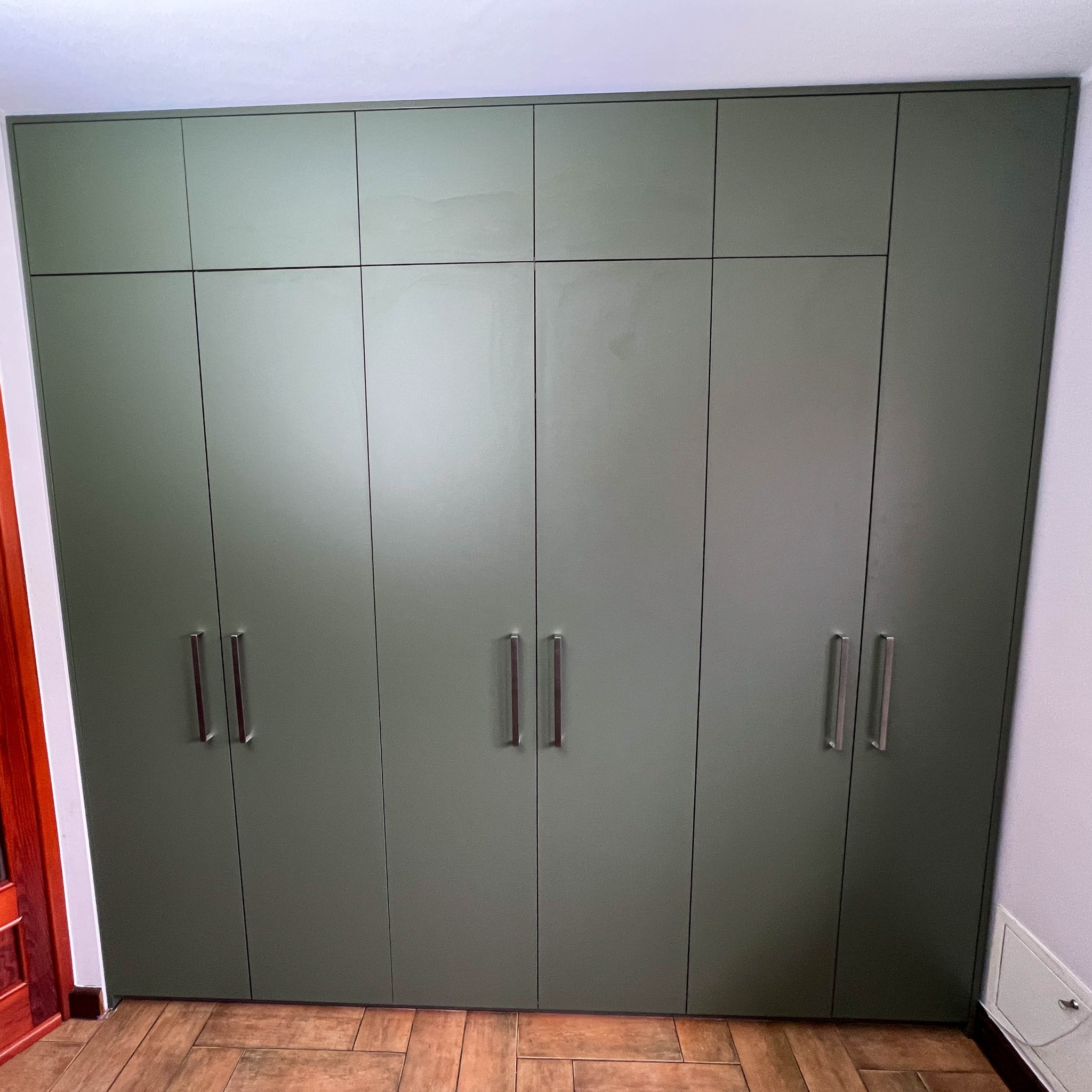 Duża, oliwkowa szafa wnękowa z metalowymi uchwytami, dopasowana do sufitu, na podłodze z płytek imitujących drewno.