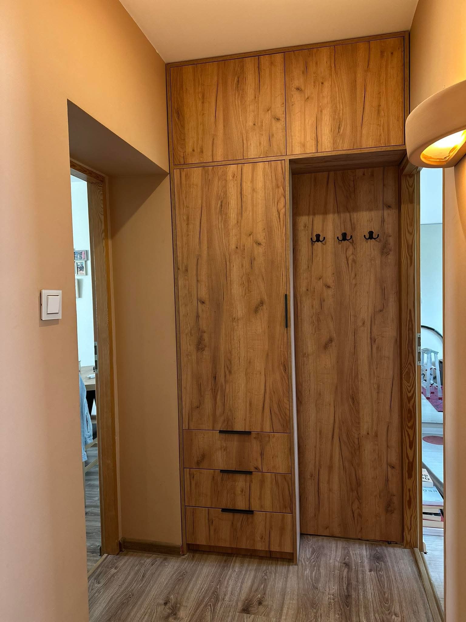 Wysoka szafa z frontami imitującymi drewno, z trzema szufladami na dole i górną szafką, wkomponowana w przedpokój z wieszakami na ścianie wnęki.