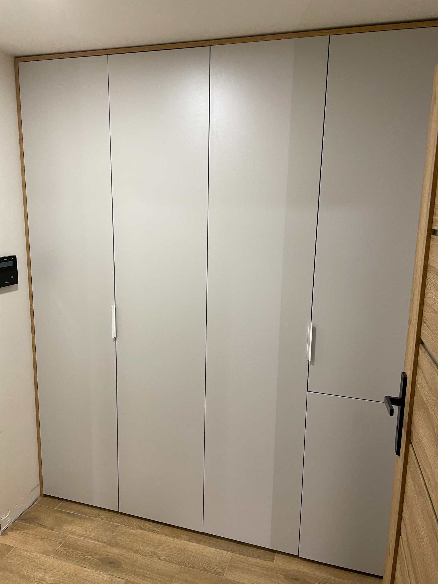 Jasnoszara, czterodrzwiowa szafa wnękowa z drewnianą ramą, minimalistyczne uchwyty, widoczna część drewnianych drzwi wejściowych.