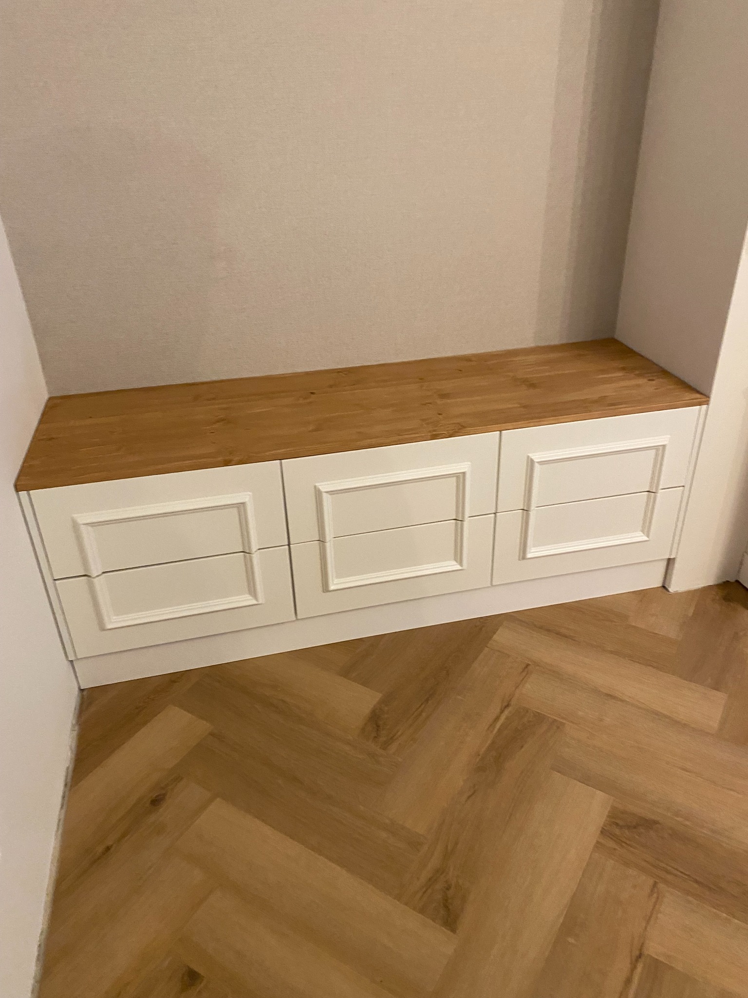 Biała szafka z trzema szufladami z dekoracyjnymi ramkami i drewnianym blatem, wkomponowana we wnękę, parkiet w jodełkę.