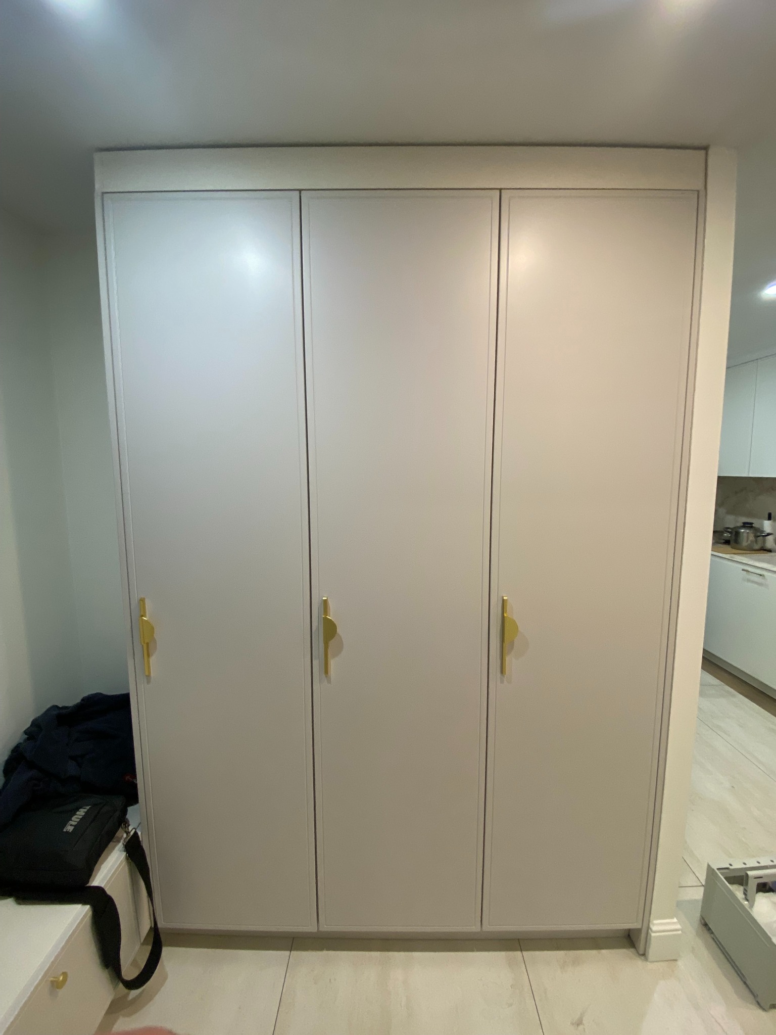 Trzydrzwiowa szafa wnękowa z jasnoszarymi frontami i złotymi uchwytami, widoczny fragment kuchni w tle.