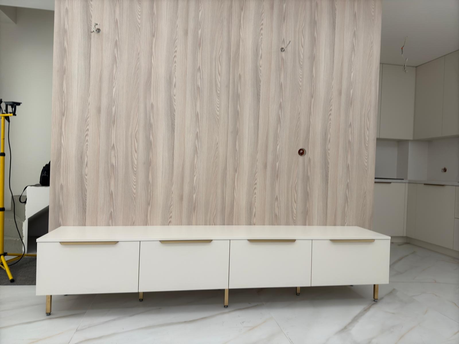 Biała szafka RTV z czterema szufladami i złotymi uchwytami na tle ściany z drewnopodobną okleiną i fragmentu białej kuchni.