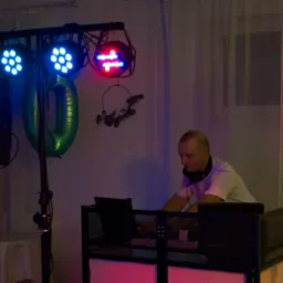 DJ w trakcie pracy, oświetlenie sceniczne z niebieskimi i czerwonymi reflektorami, głośniki po bokach. W tle dekoracje i zasłony.