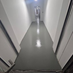 „Azazeli21 - Nowa, błyszcząca, szara wylewka w wąskim korytarzu, widok z perspektywy otwartych drzwi. Białe ściany i sufit, lampa sufitowa. Świeżo wykonana podłoga.