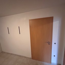 „Azazeli21 - Biała ściana z dwiema czarnymi kreskami, brązowe drzwi z klamką, pralka i płytki podłogowe w pomieszczeniu. Widoczne gniazdka elektryczne na ścianie.