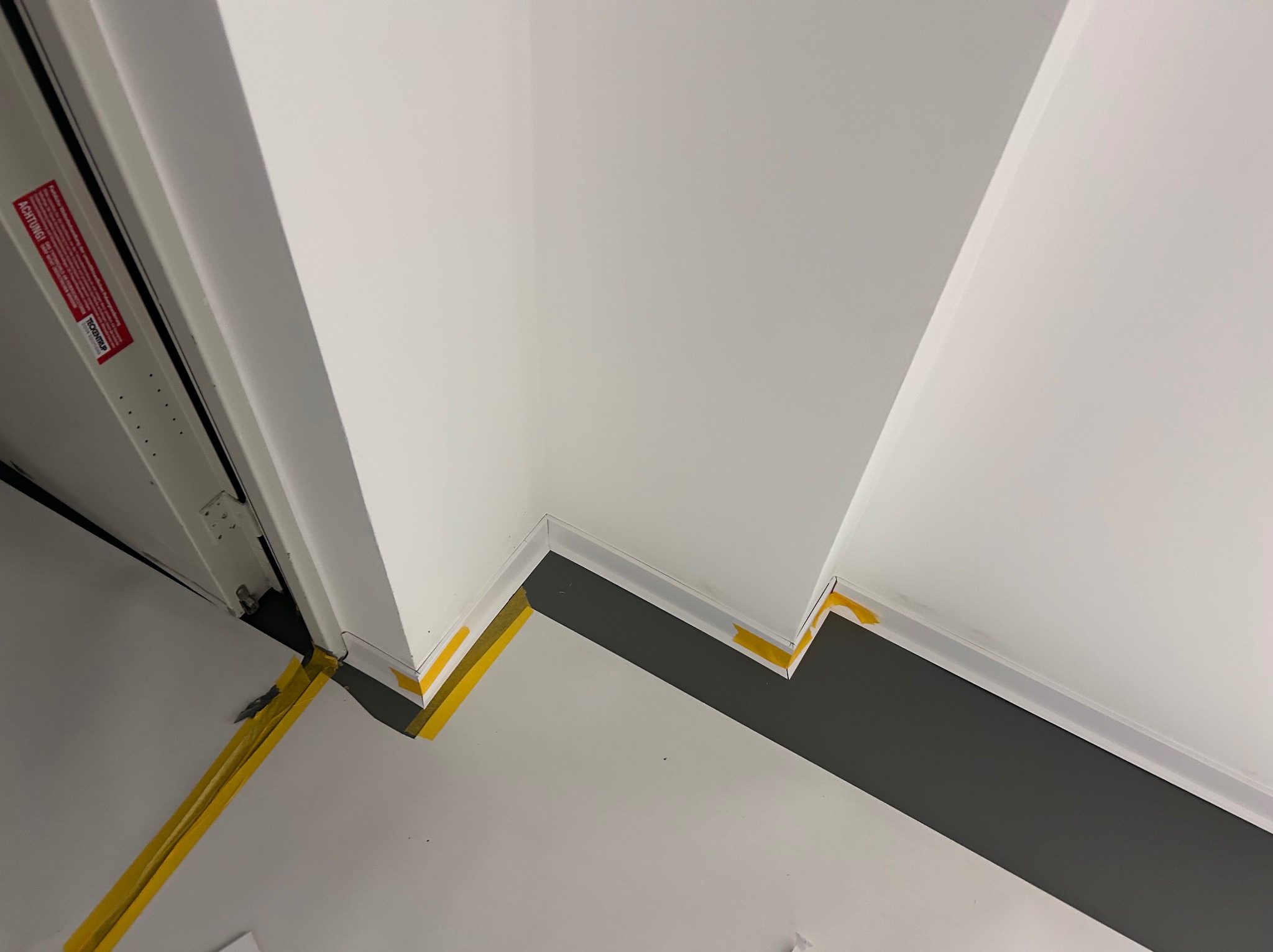 Świeżo zamontowana listwa przypodłogowa w narożniku, zabezpieczona żółtą taśmą malarską, na styku białej ściany i szarej podłogi. Widoczne detale wykończenia.