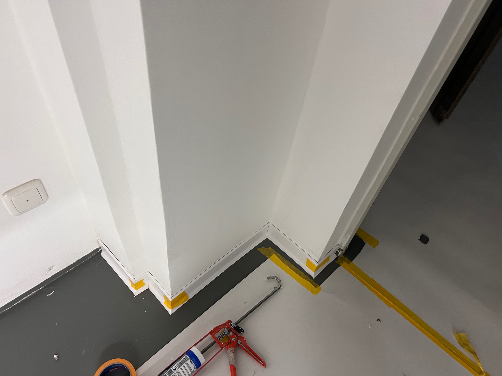 Białe listwy przypodłogowe zabezpieczone żółtą taśmą malarską, narożniki wykończone silikonem. Widoczny aplikator do silikonu i fragment podłogi w szarym kolorze.