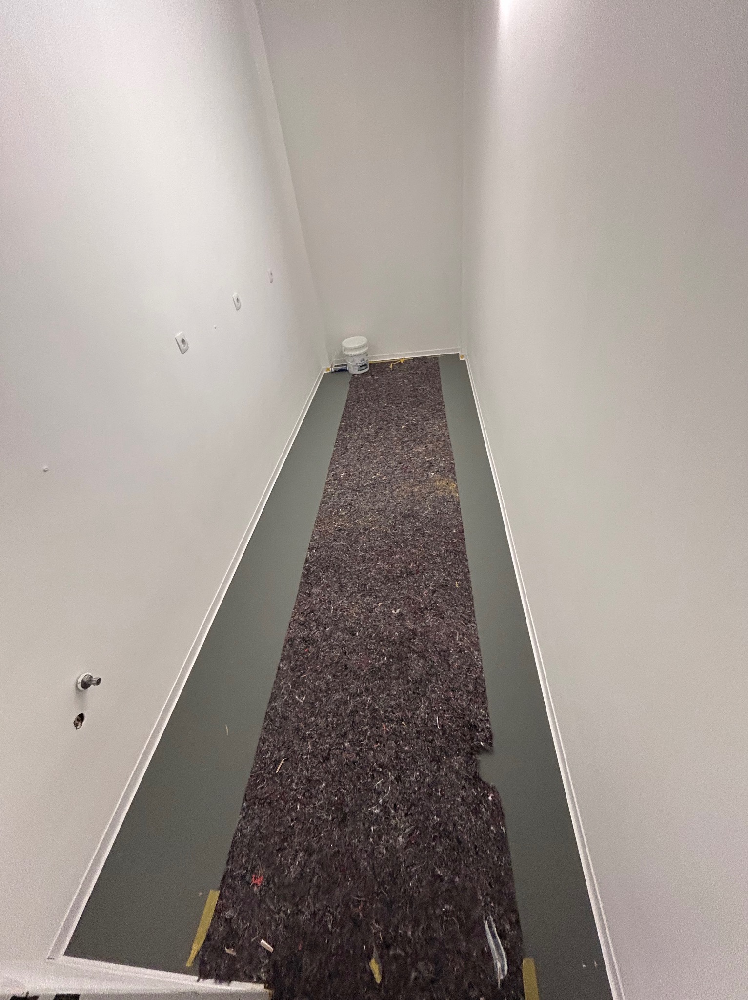 Przygotowanie podłoża w korytarzu: podkład pod panele na szarej wylewce, białe ściany z gniazdkami, wiadro farby w tle. Widok z góry.