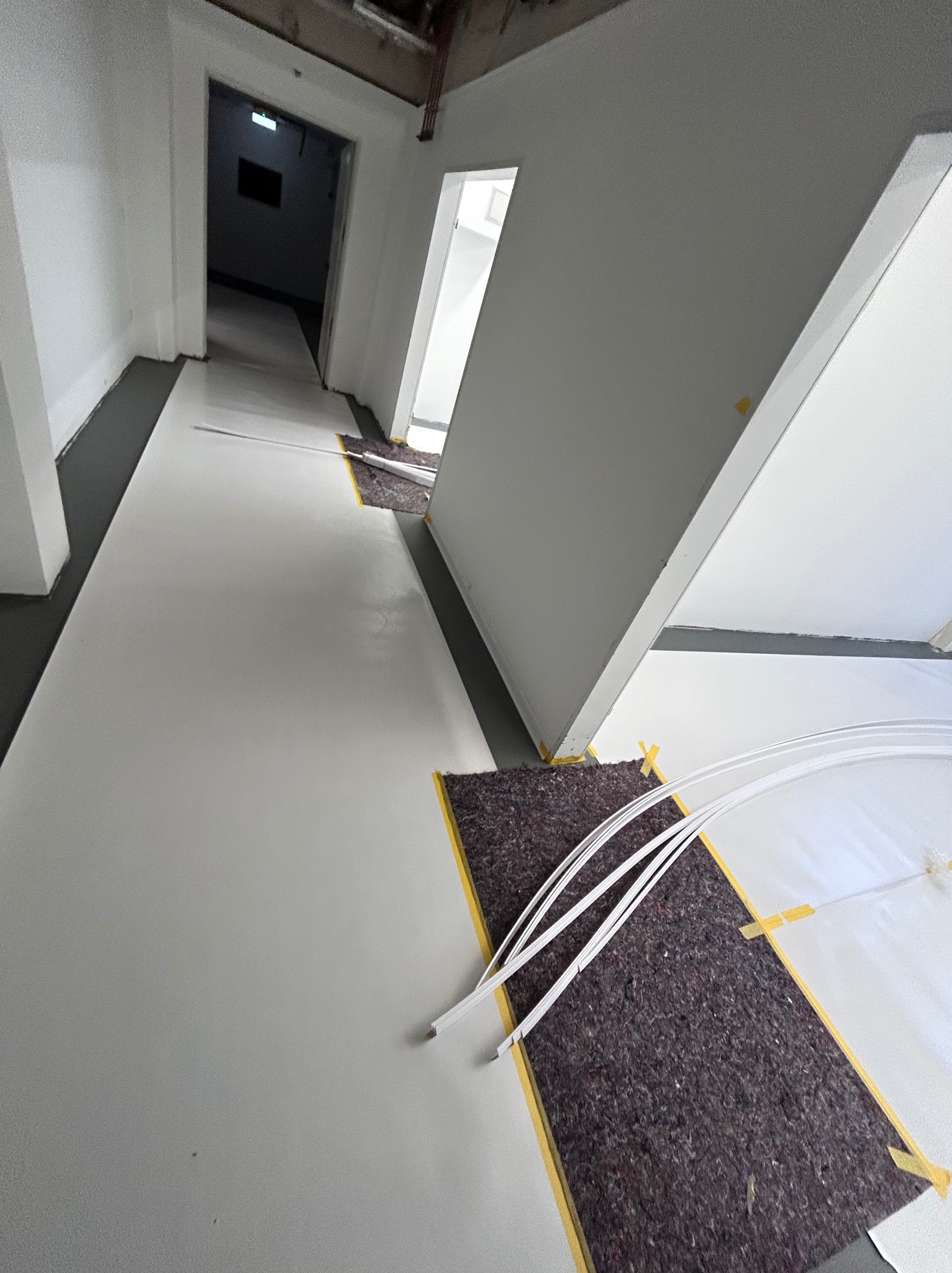 Biała, gładka posadzka w korytarzu podczas prac wykończeniowych, zabezpieczona folią i tekturą falistą. Widoczne kable i taśmy malarskie.