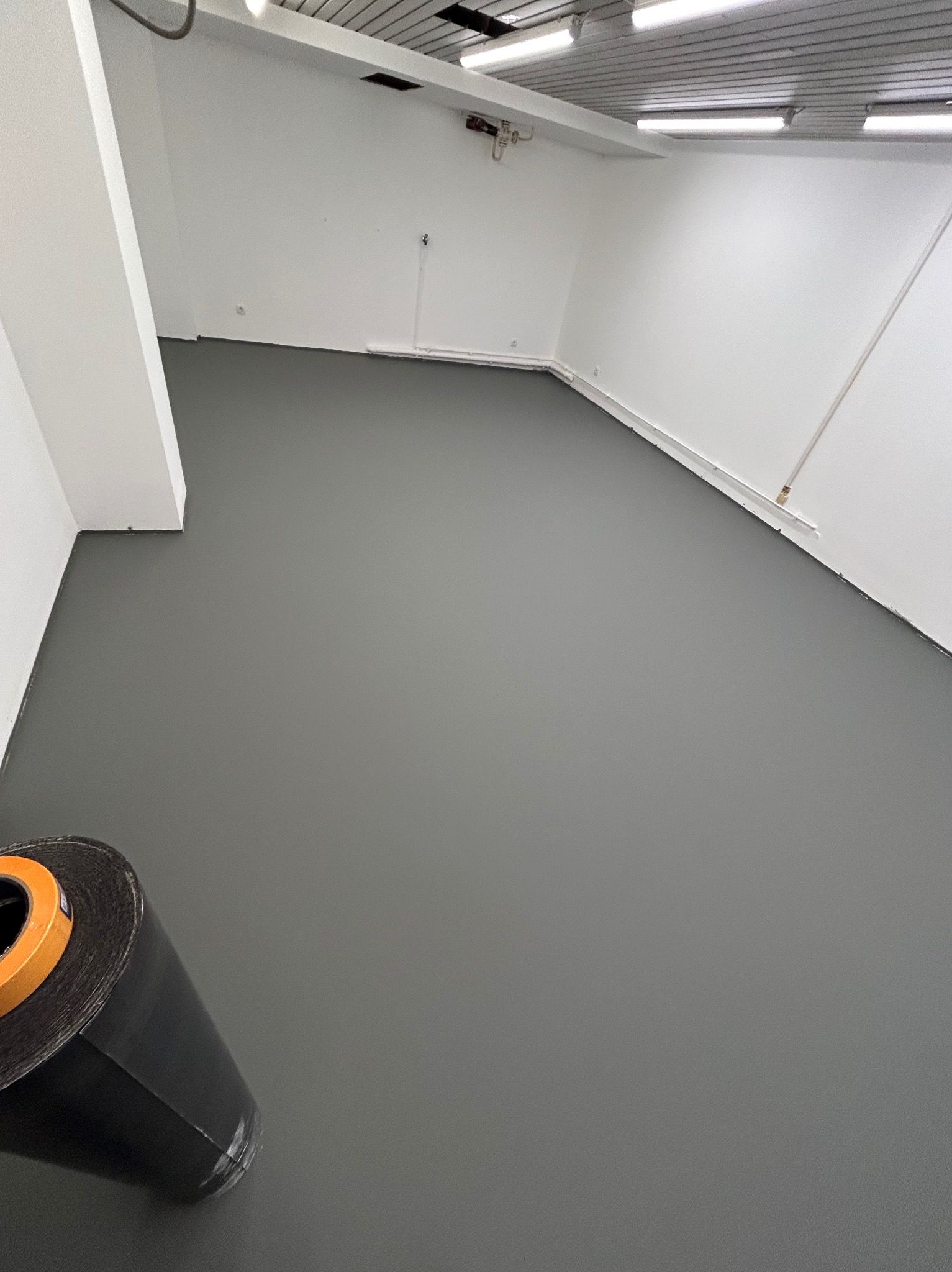 Szara, gładka posadzka w pomieszczeniu biurowym po wylaniu wylewki samopoziomującej. W rogu widoczna rolka folii. Białe ściany i sufit z oświetleniem.