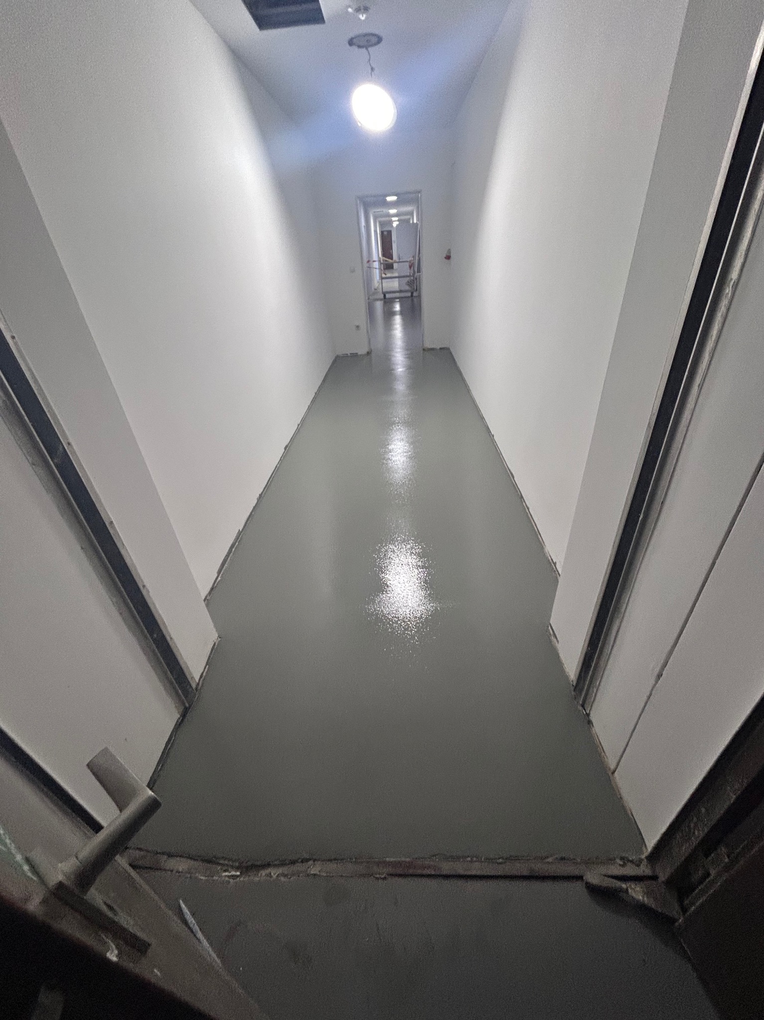 Nowa, błyszcząca, szara wylewka w wąskim korytarzu, widok z perspektywy otwartych drzwi. Białe ściany i sufit, lampa sufitowa. Świeżo wykonana podłoga.