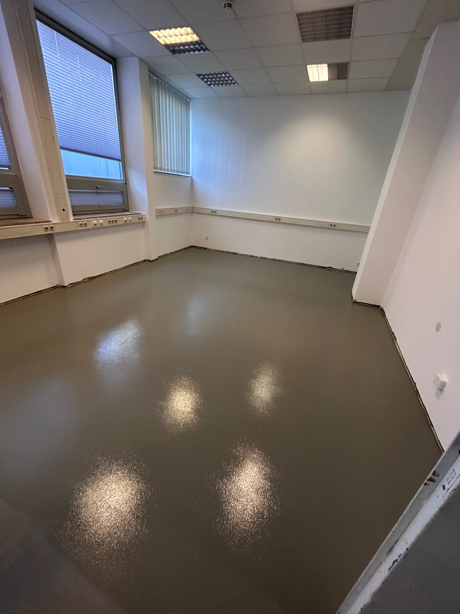 Świeżo wylana, lśniąca posadzka w jasnym biurze. Widoczne okna z żaluzjami, białe ściany z listwami przypodłogowymi i fragment drabiny malarskiej w prawym dolnym rogu.
