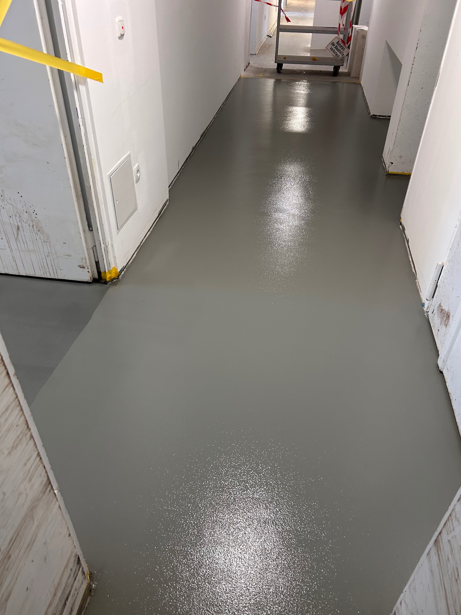 Świeżo wylana, błyszcząca, szara posadzka epoksydowa w korytarzu biurowym, zabezpieczona żółtą taśmą przy drzwiach i ścianach, z widocznym wózkiem transportowym w tle.