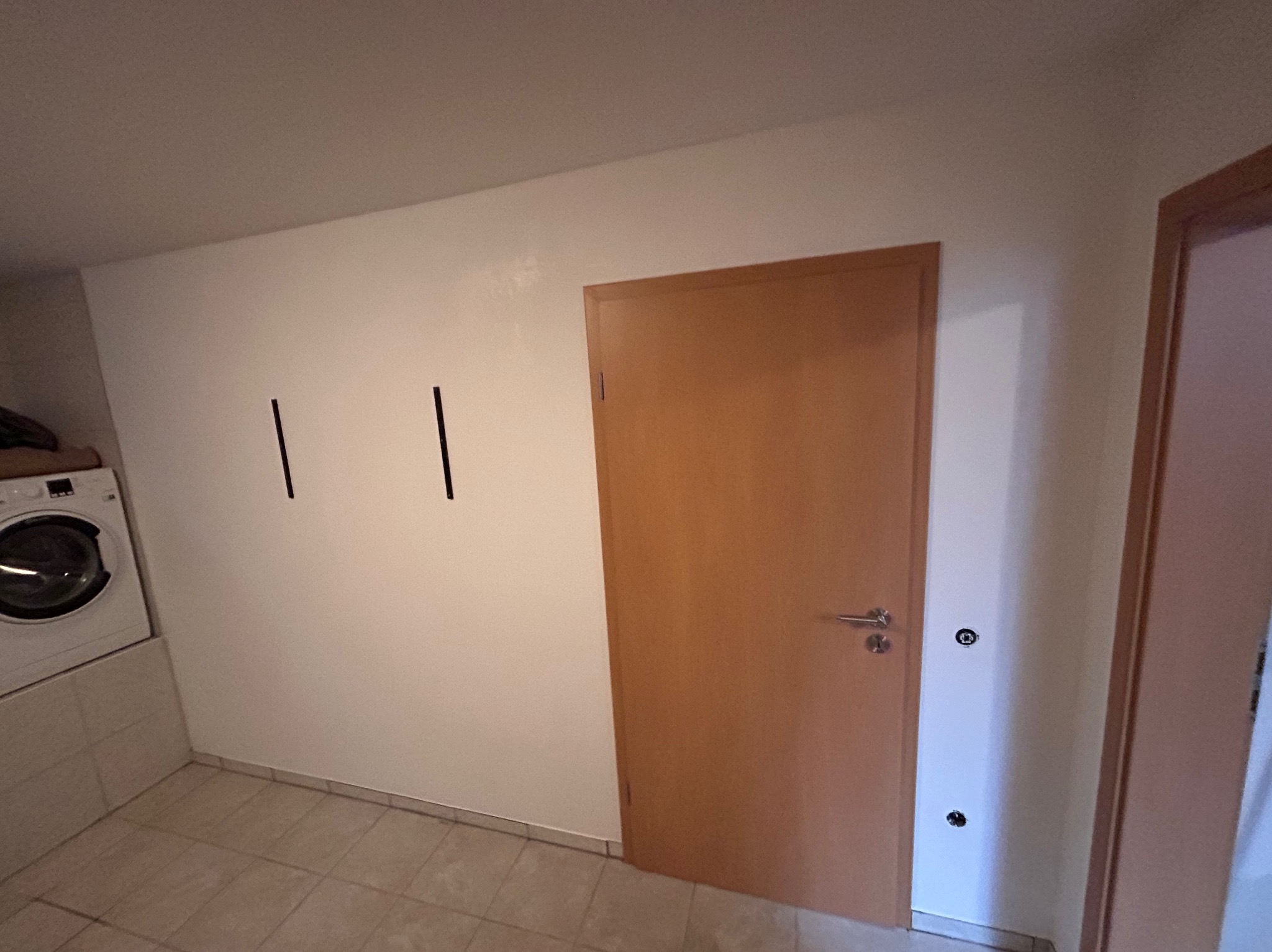 Biała ściana z dwiema czarnymi kreskami, brązowe drzwi z klamką, pralka i płytki podłogowe w pomieszczeniu. Widoczne gniazdka elektryczne na ścianie.
