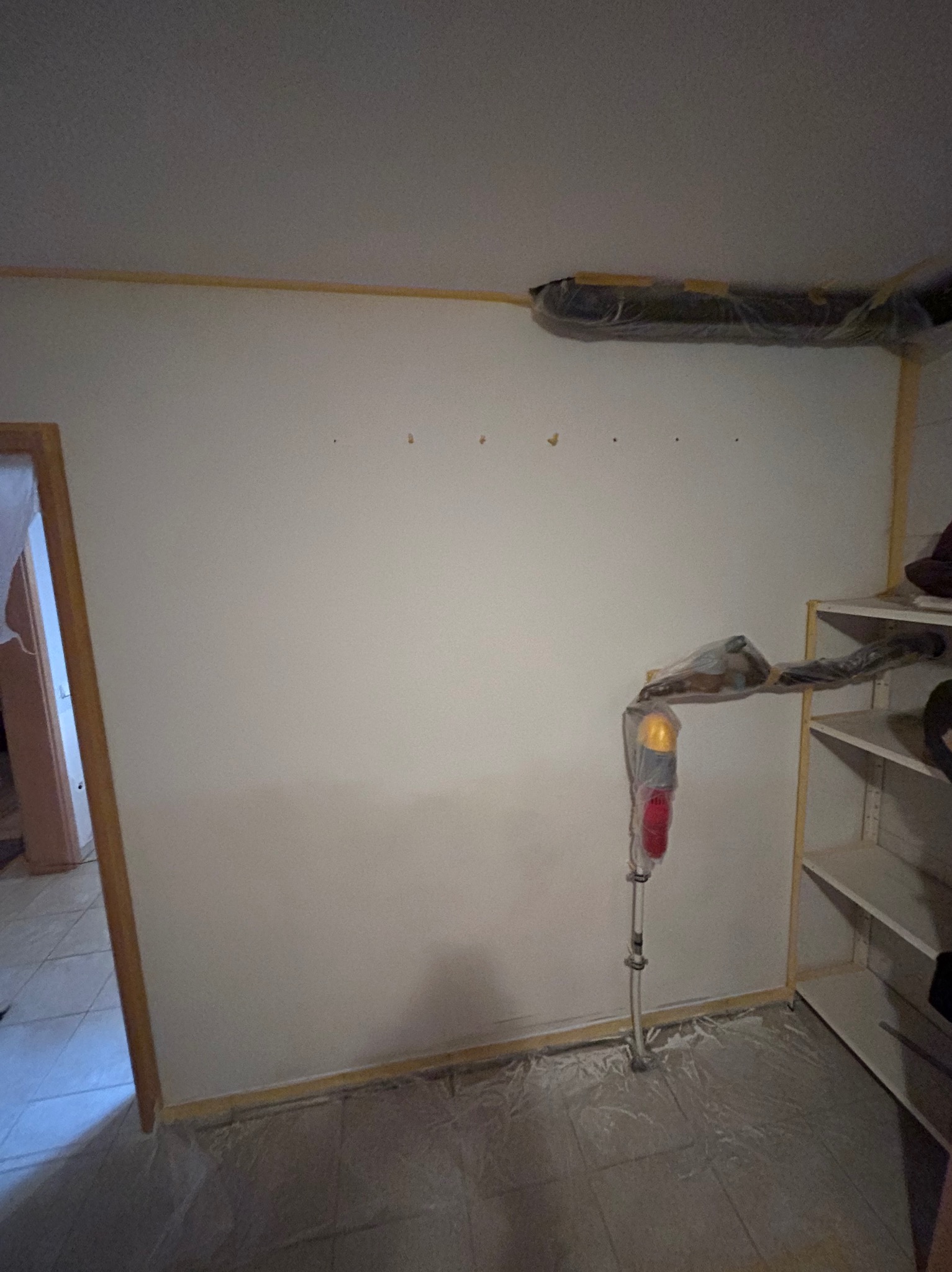 Przygotowana ściana do malowania z zaklejonymi listwami i podłogą folią ochronną. Widoczna drabina malarska i regał z półkami.