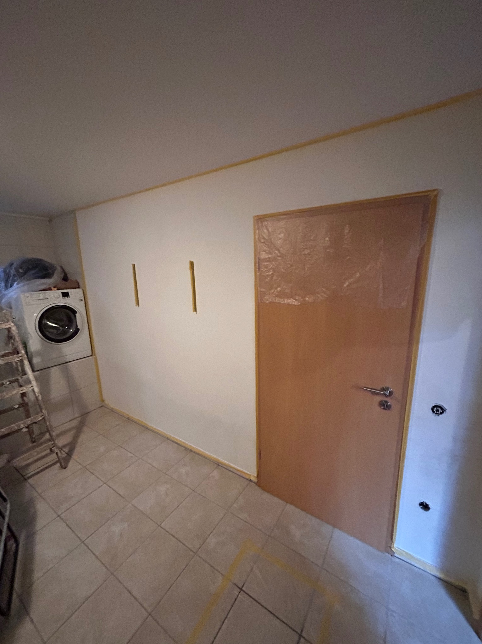 Przygotowania do malowania: zabezpieczona folią drzwi, taśma malarska na ścianach i podłodze w pomieszczeniu z pralką i drabinką.