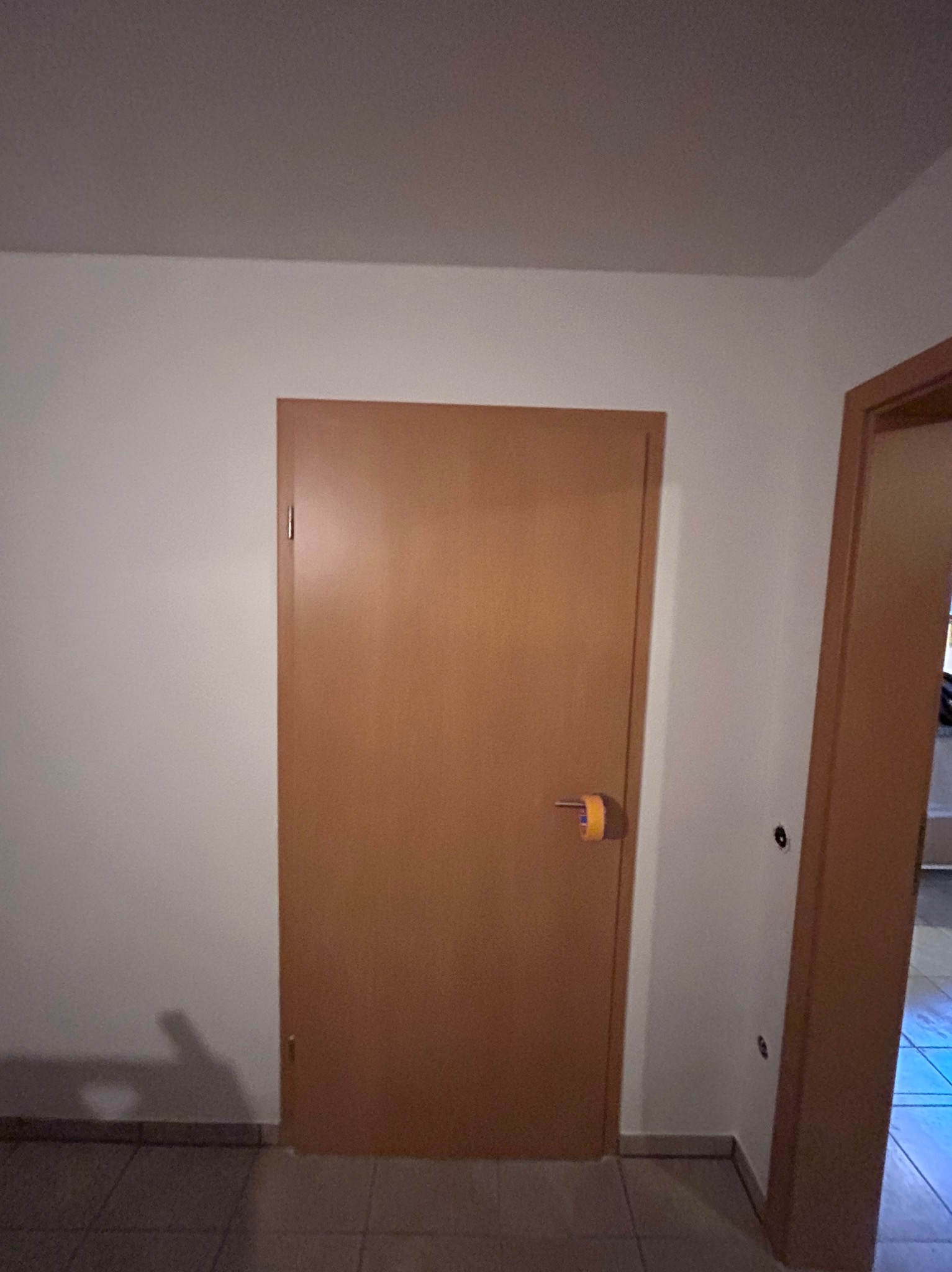 Wnętrze: pomalowana ściana w odcieniach bieli, brązowe drzwi z taśmą malarską na klamce i fragment drugich drzwi. Minimalistyczny styl, brak dekoracji.