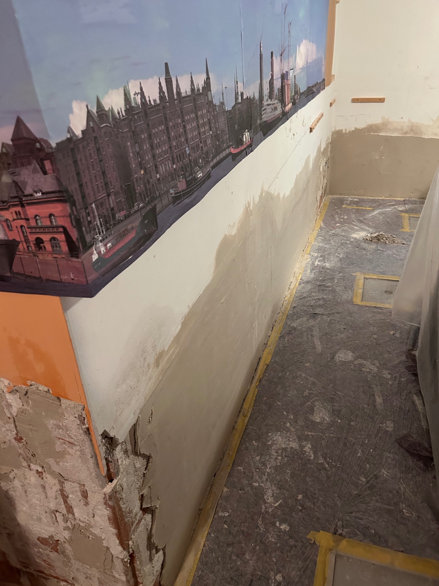 Ściana w trakcie szpachlowania, z widoczną tapetą z motywem miejskim i fragmentem starego muru. Przygotowanie do malowania, widoczne zabezpieczenia podłogi.