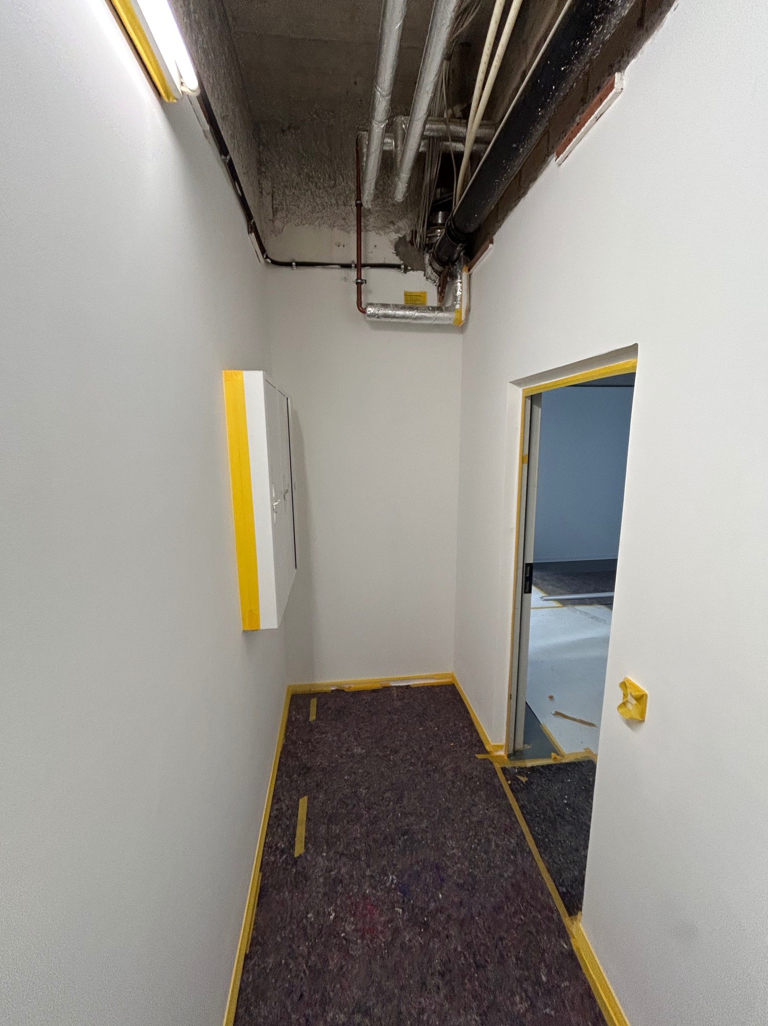 Korytarz w trakcie malowania: świeżo pomalowane ściany na szaro, zabezpieczone taśmą malarską, widoczne rury pod sufitem i szafka elektryczna. Etap prac wykończeniowych.