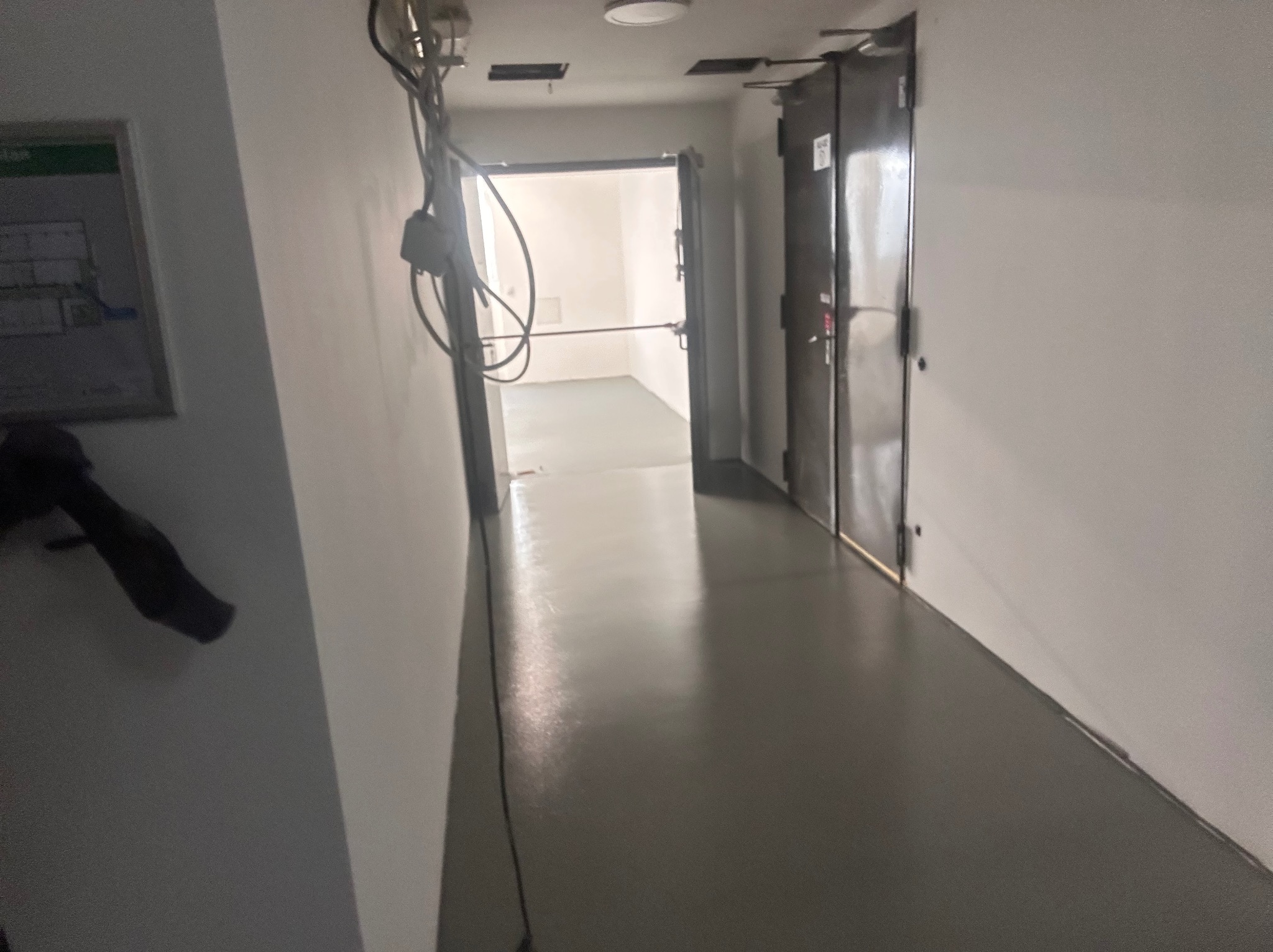 Korytarz biurowy z gładką, szarą podłogą i białymi ścianami. Widoczne kable wystające ze ściany, drzwi po prawej i plan budynku na ścianie po lewej.