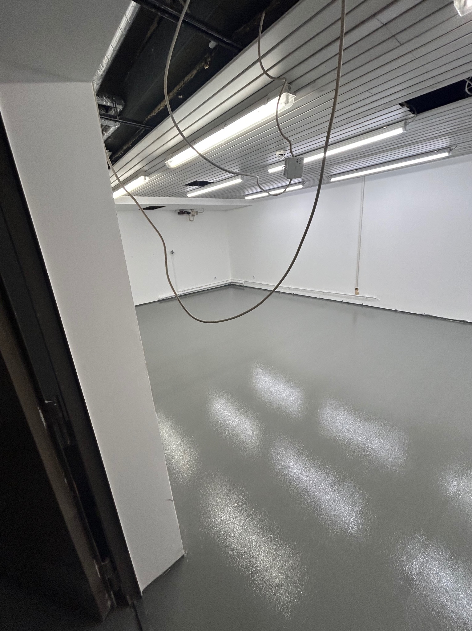 Wykończone wnętrze biurowe z szarą, błyszczącą posadzką, białymi ścianami i widocznymi przewodami pod sufitem. Oświetlenie jarzeniowe. Widok zza otwartych drzwi.