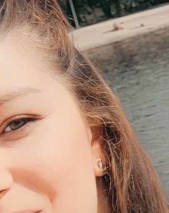 Portret kobiety z bliska, z widocznym okiem, uchem z kolczykiem i fragmentem włosów. W tle woda i plaża z leżącą osobą.