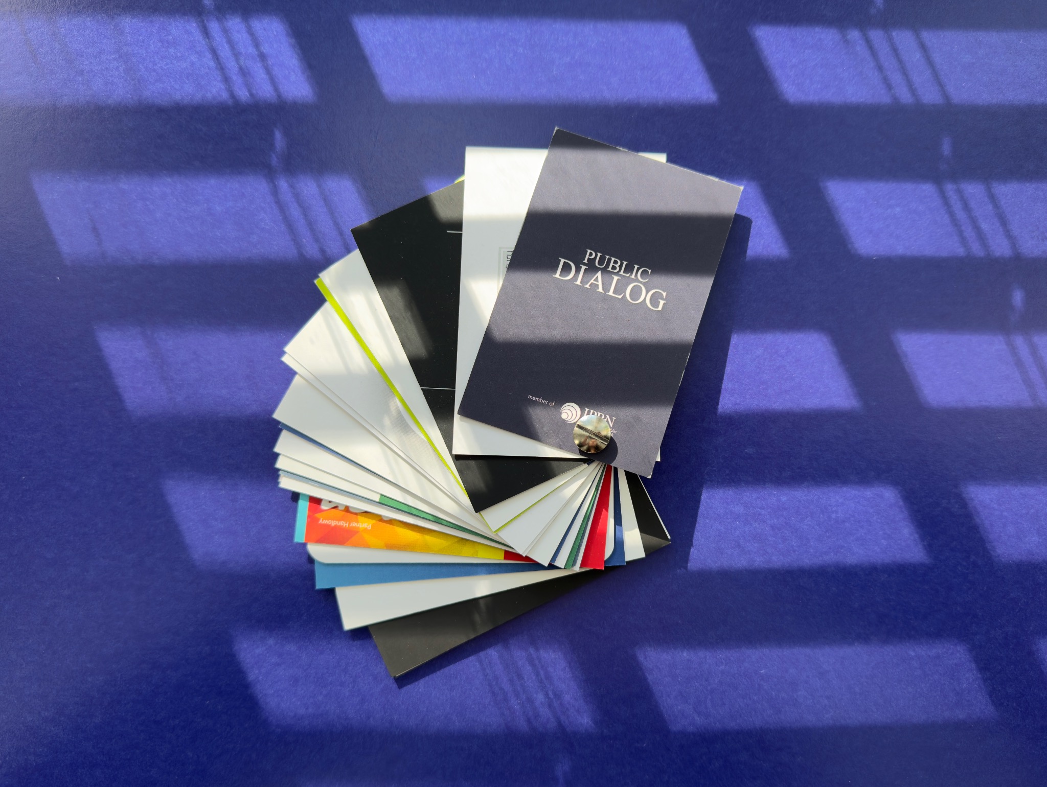 Kaskada różnorodnych, gotowych wizytówek z napisem 'PUBLIC DIALOG' na wierzchu, ułożonych wachlarzowo na fioletowym tle z cieniem okiennym.