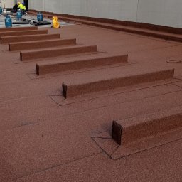 PBN INVEST PLUS SPÓŁKA Z OGRANICZONĄ ODPOWIEDZIALNOŚCIĄ - Widok z góry na brązową papę termozgrzewalną na dachu z wystającymi elementami, w tle pracownicy z butlami gazowymi.