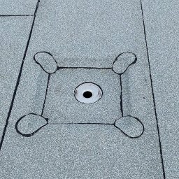 PBN INVEST PLUS SPÓŁKA Z OGRANICZONĄ ODPOWIEDZIALNOŚCIĄ - Szary, płaski dach pokryty papą z centralnie umieszczonym odpływem wody deszczowej obrobionym blachą i uszczelnionym czarnym kitem dekarskim.