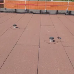 Widok z góry na płaski dach pokryty papą w kolorze ceglastym, z widocznymi dwoma odpowietrznikami i elementami zabezpieczeń na krawędzi dachu.