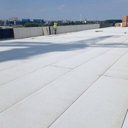 PBN INVEST PLUS SPÓŁKA Z OGRANICZONĄ ODPOWIEDZIALNOŚCIĄ - Pracownik w kamizelce odblaskowej na rozległym, płaskim dachu pokrytym jasną membraną, z widocznymi płytami izolacyjnymi i elementami konstrukcyjnymi, na tle panoramy miasta w oddali.
