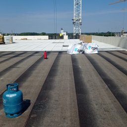 PBN INVEST PLUS SPÓŁKA Z OGRANICZONĄ ODPOWIEDZIALNOŚCIĄ - Widok z góry na płaski dach w trakcie prac, z widocznymi pasami papy termozgrzewalnej, butlą gazową, gaśnicą, workami z materiałem izolacyjnym oraz pracownikiem w oddali i żurawiem w tle.