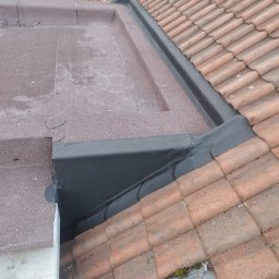 PBN INVEST PLUS SPÓŁKA Z OGRANICZONĄ ODPOWIEDZIALNOŚCIĄ - Połączenie dachu płaskiego pokrytego papą z dachem skośnym z dachówki ceramicznej, z widocznymi obróbkami blacharskimi w kolorze czarnym.