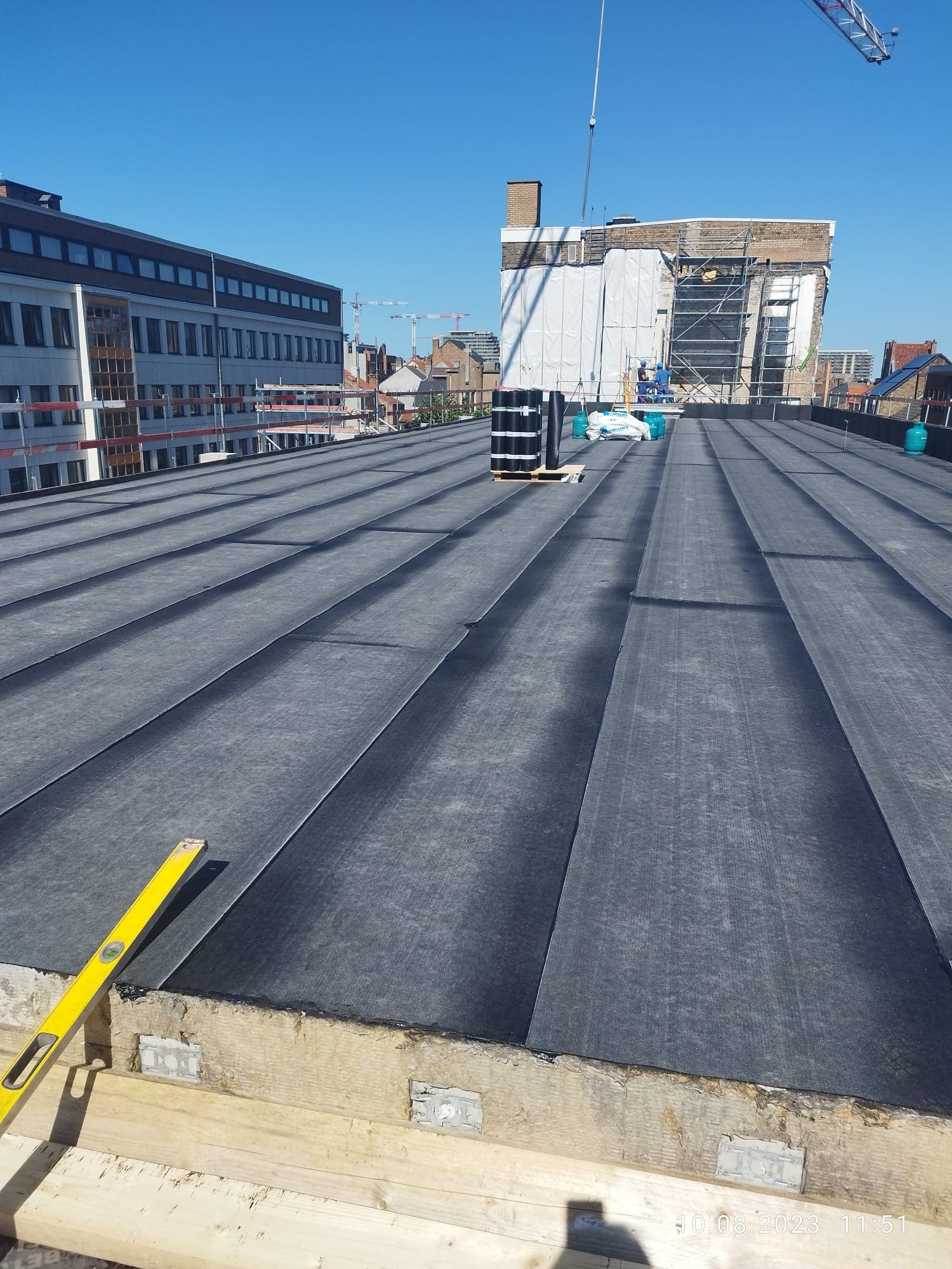 Nowe pokrycie dachu płaskiego z papy termozgrzewalnej, widoczny poziomica, rolki papy, butle gazowe, w tle budynki i żuraw budowlany.