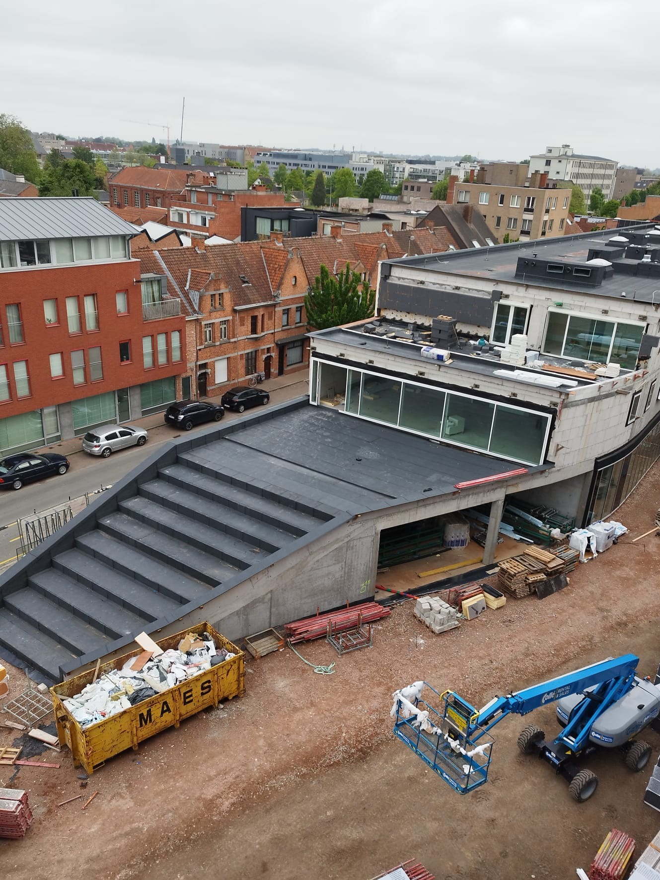 Widok z góry na budynek z płaskim dachem w trakcie budowy, z widocznymi schodami prowadzącymi na dach, kontenerem na śmieci budowlane i podnośnikiem koszowym na placu budowy