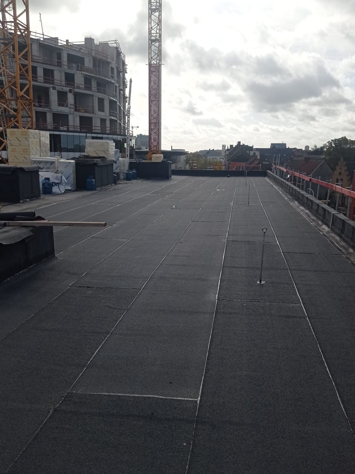 Wykończony płaski dach pokryty papą termozgrzewalną z widocznymi metalowymi uchwytami, w tle budynek w budowie i żuraw budowlany.
