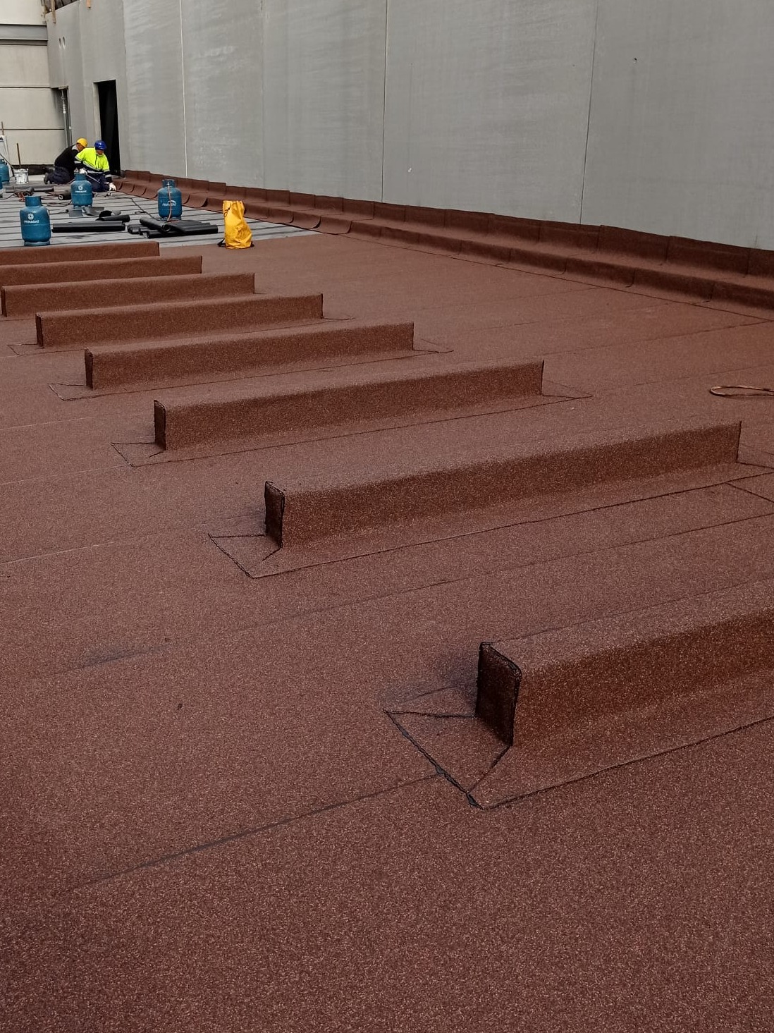 Widok z góry na brązową papę termozgrzewalną na dachu z wystającymi elementami, w tle pracownicy z butlami gazowymi.