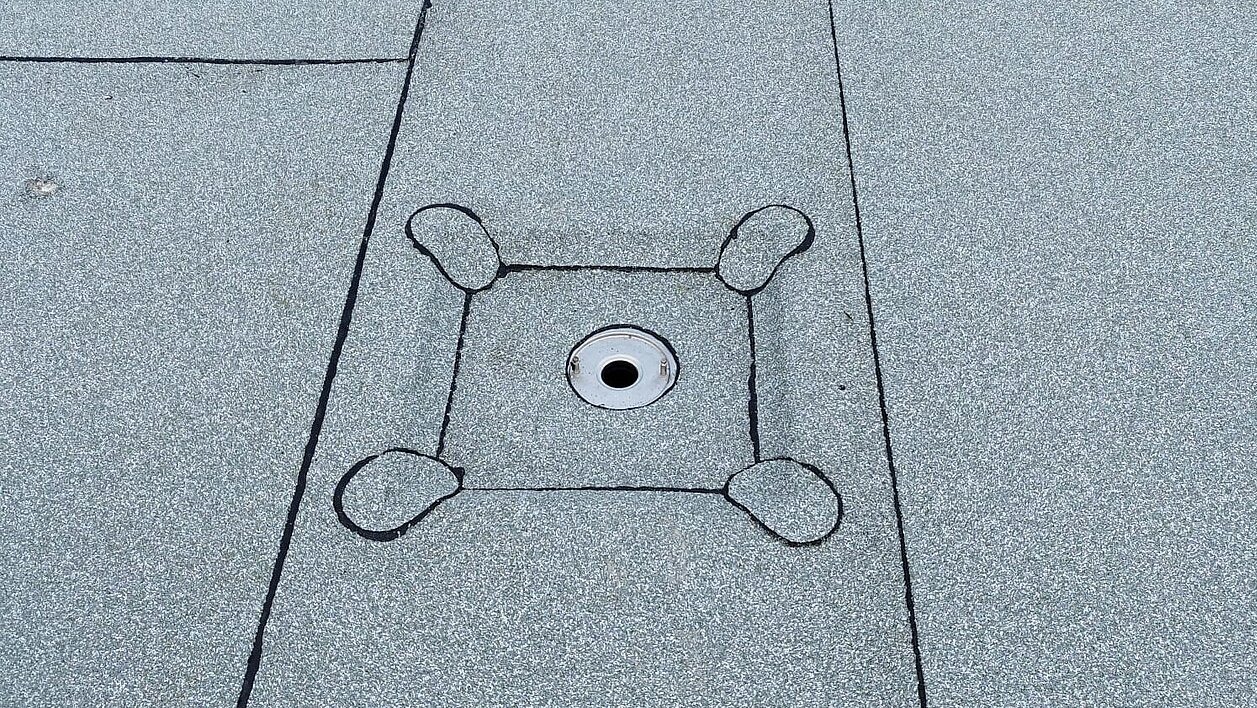 Szary, płaski dach pokryty papą z centralnie umieszczonym odpływem wody deszczowej obrobionym blachą i uszczelnionym czarnym kitem dekarskim.