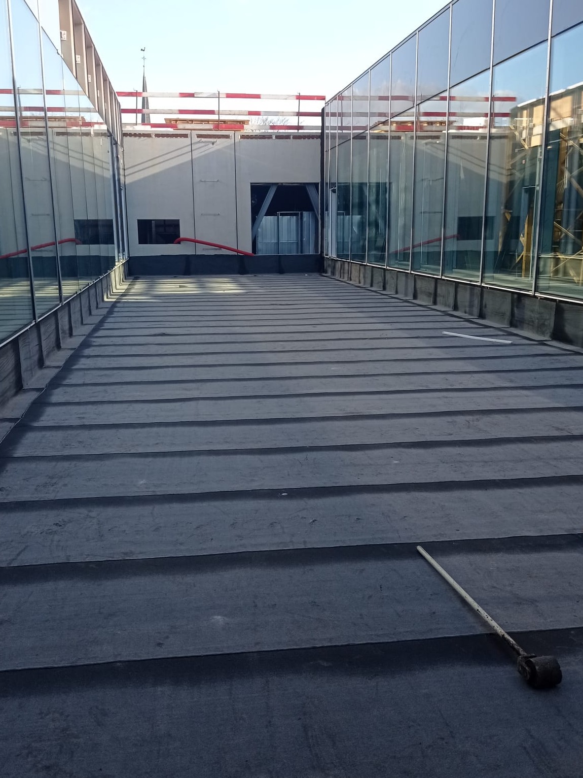 Płaski dach pokryty papą termozgrzewalną z widocznym wałkiem dekarskim, obok ściany z przeszkleniami.