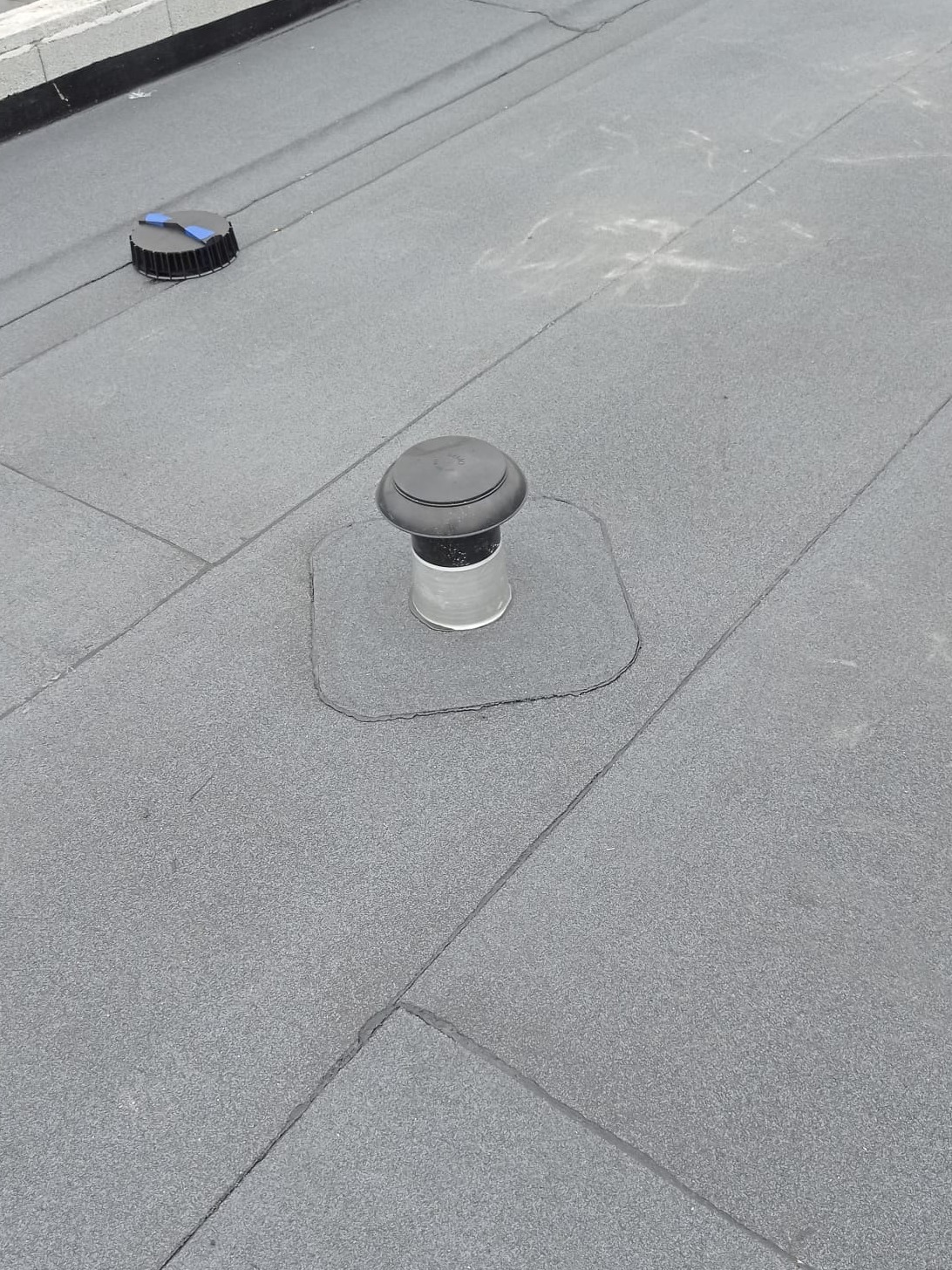 Szary, płaski dach pokryty papą z dwoma kominkami wentylacyjnymi, jeden z niebieskim elementem zabezpieczającym.