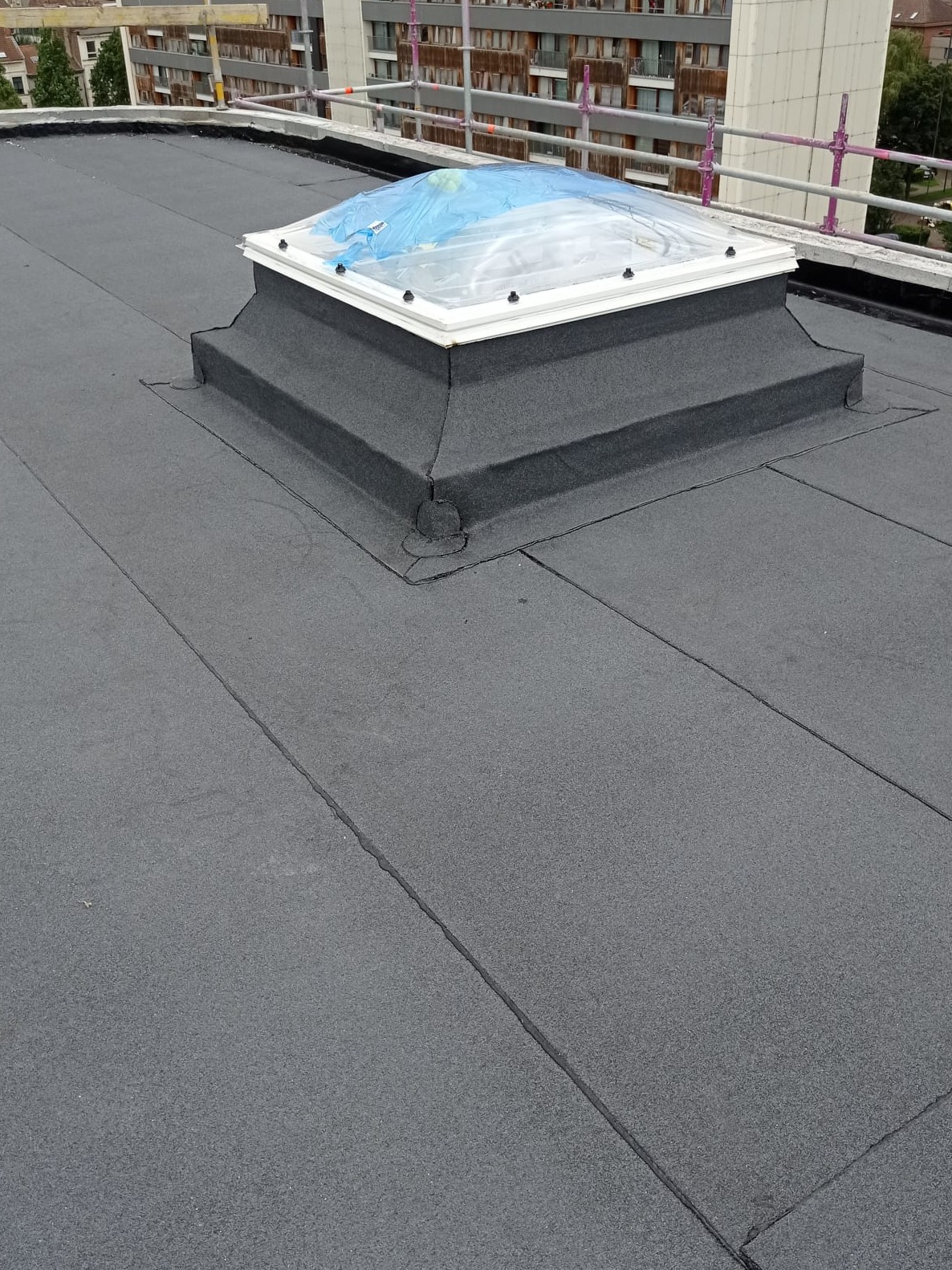 Płaski dach pokryty czarną papą z zamontowanym świetlikiem dachowym zabezpieczonym folią, widok z góry.